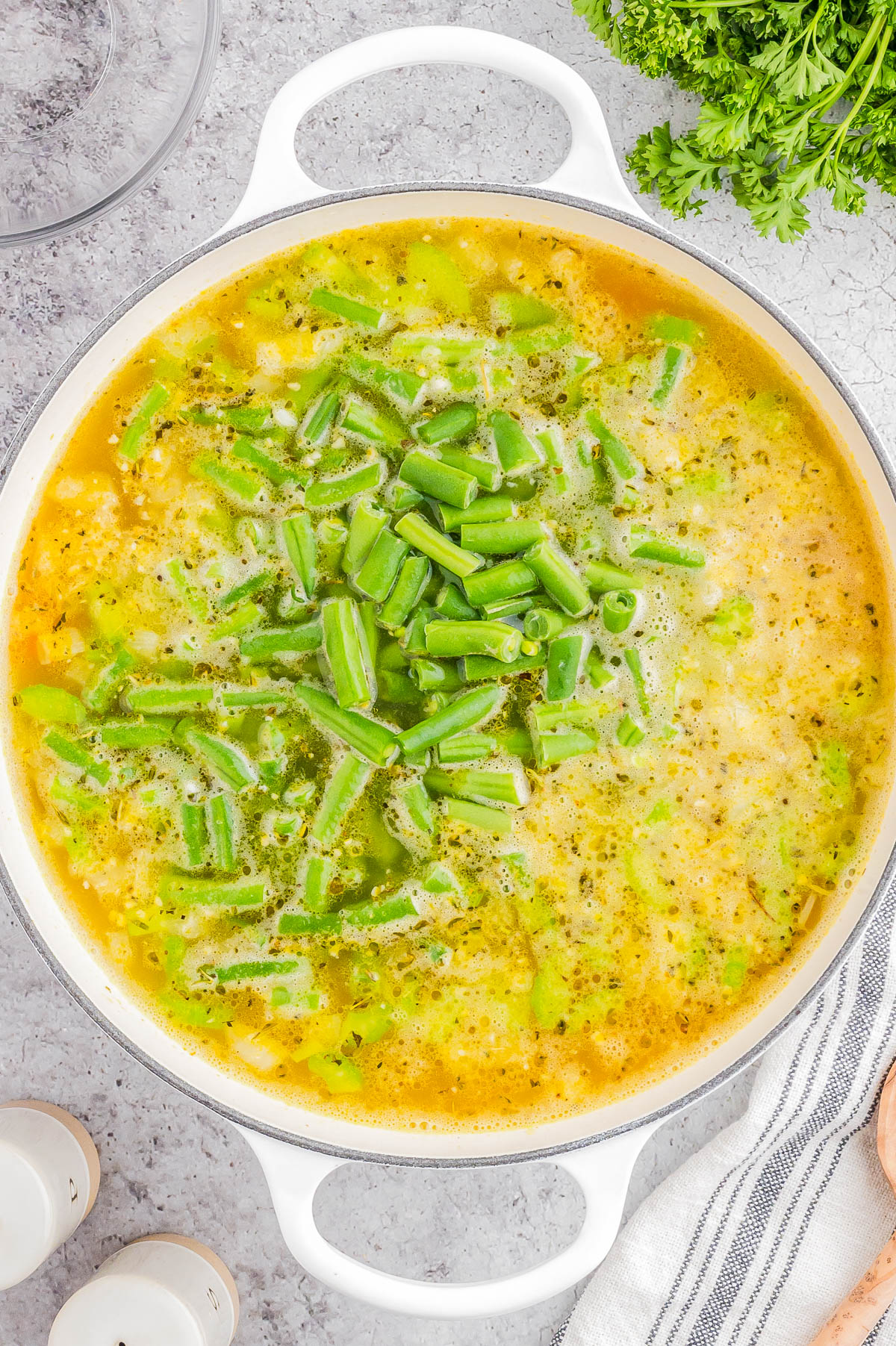 A white pot filled with a yellow-green soup topped with chopped green beans, sitting on a gray countertop with herbs, salt and pepper shakers, and a striped towel nearby.