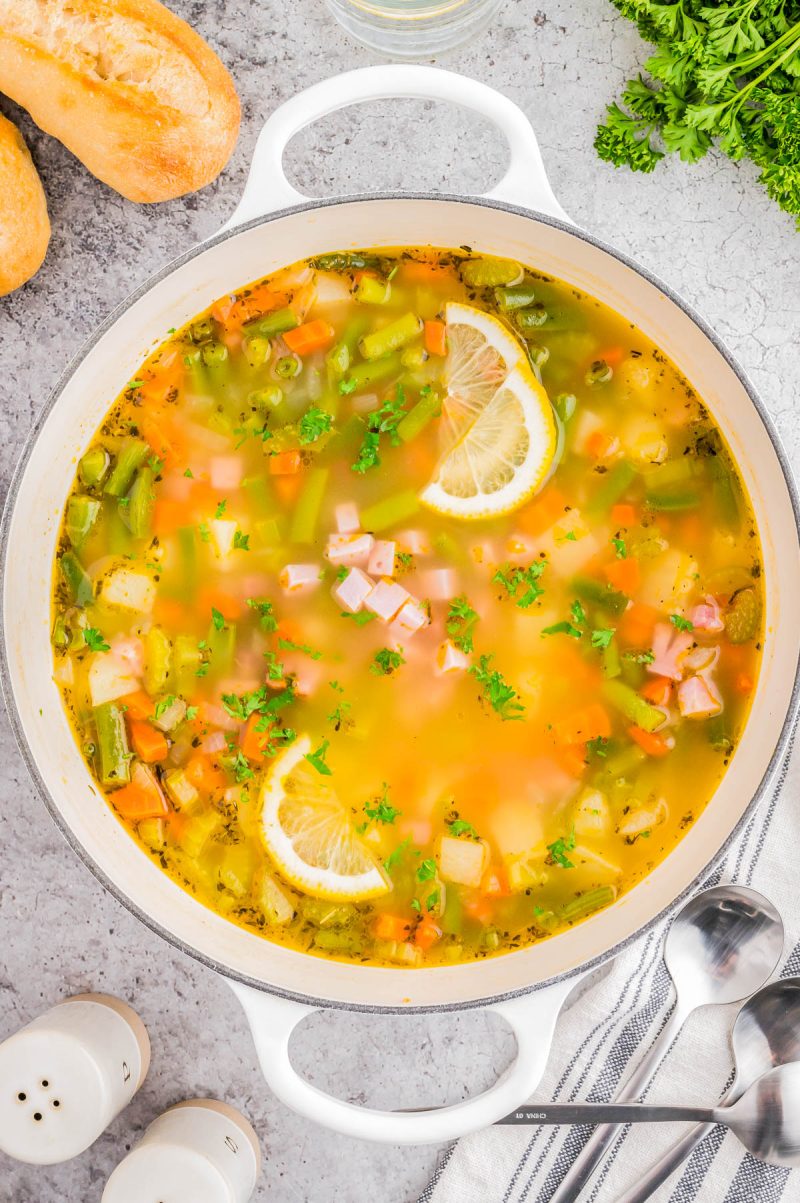 A pot of vegetable soup with diced ham, carrots, potatoes, green beans, and parsley, garnished with lemon slices, sits on a gray countertop beside bread and fresh herbs.