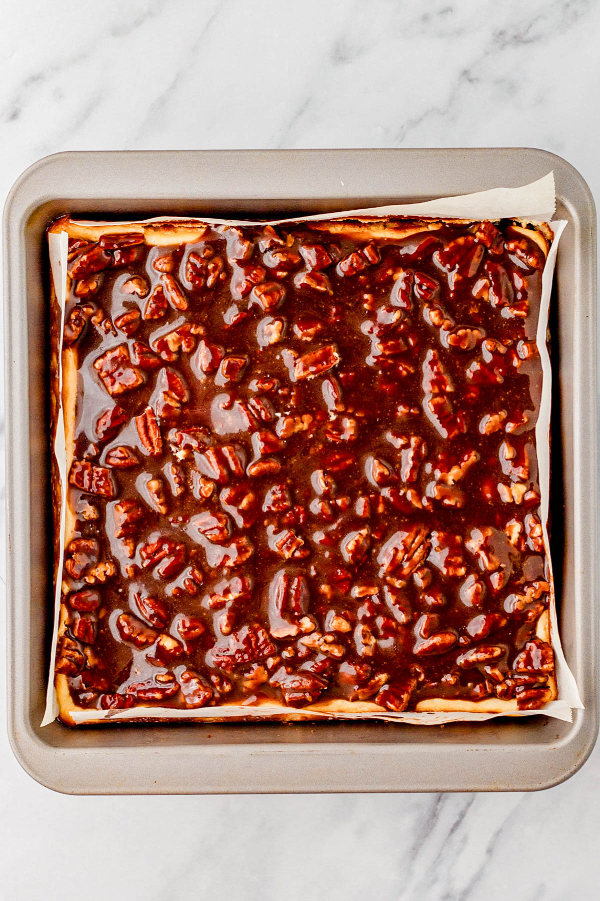 A square baking pan filled with unbaked pecan pie bars, showing a nutty, glossy brown filling spread over a crust on parchment paper.