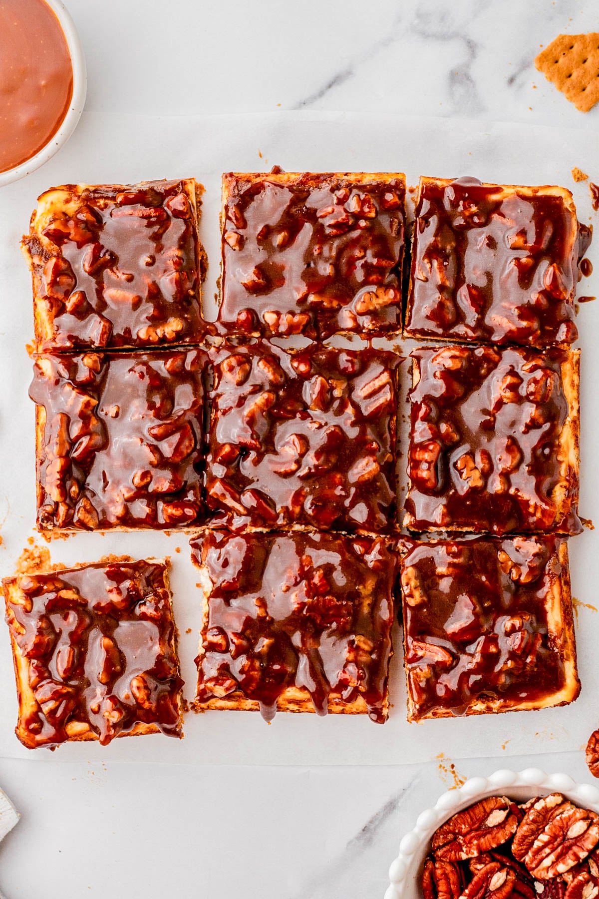 Nine square cheesecake bars topped with glossy caramel pecan sauce are arranged on a white surface; pecans and caramel are visible nearby.