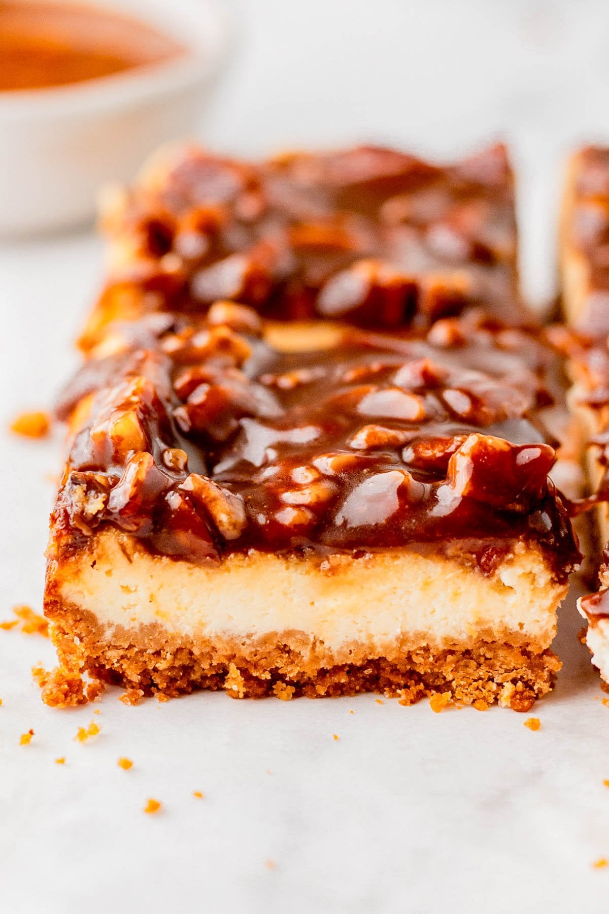 A close-up of a dessert bar with a crumbly crust, creamy middle layer, and glossy pecan topping on a light surface.