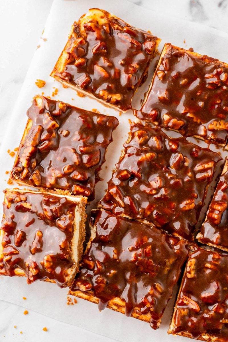 Nine square bars topped with a glossy caramel pecan glaze are arranged on a parchment-lined surface.