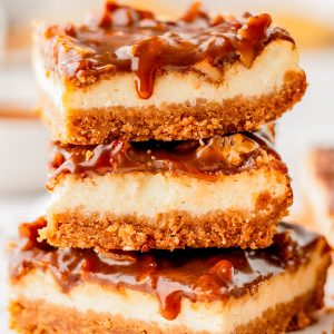 Three caramel cheesecake bars with a graham cracker crust are stacked on top of each other, with caramel sauce dripping down the sides.