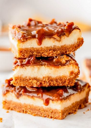 Three caramel cheesecake bars with a graham cracker crust are stacked on top of each other, with caramel sauce dripping down the sides.