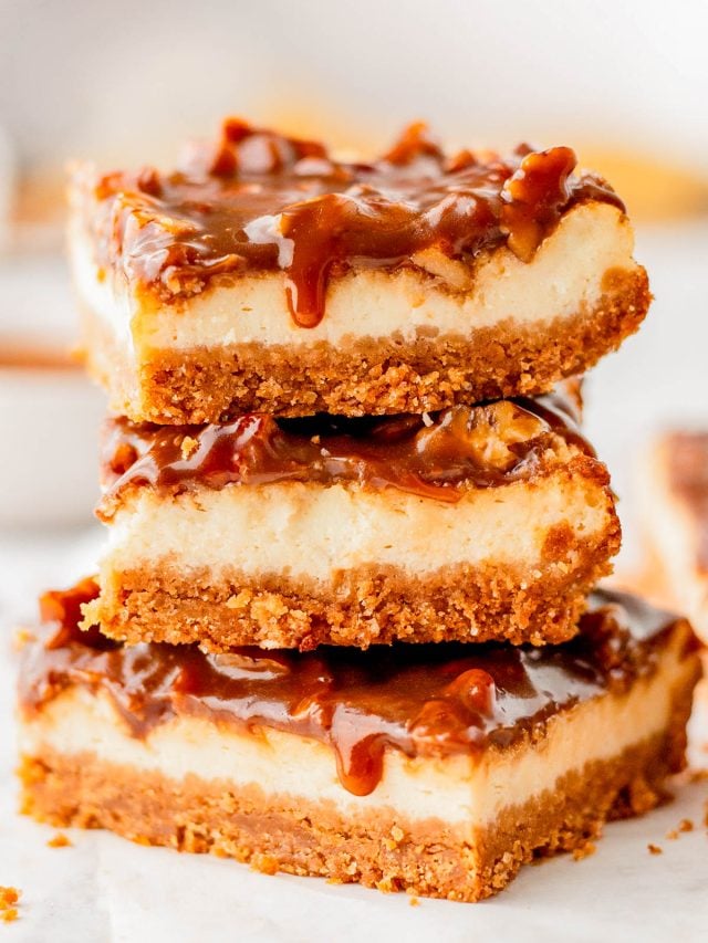 Three caramel cheesecake bars with a graham cracker crust are stacked on top of each other, with caramel sauce dripping down the sides.
