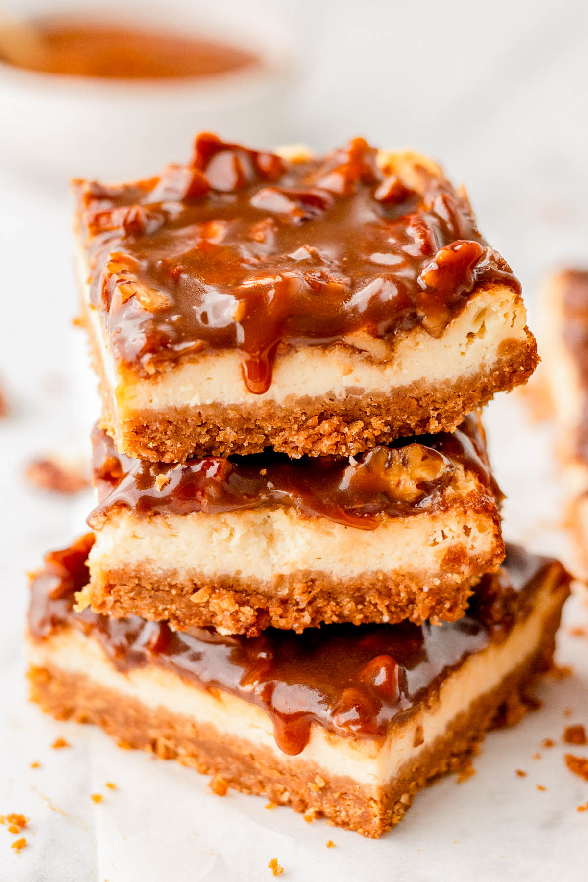 Three caramel pecan cheesecake bars are stacked on top of each other, showing layers of graham cracker crust, creamy cheesecake, and caramel pecan topping.