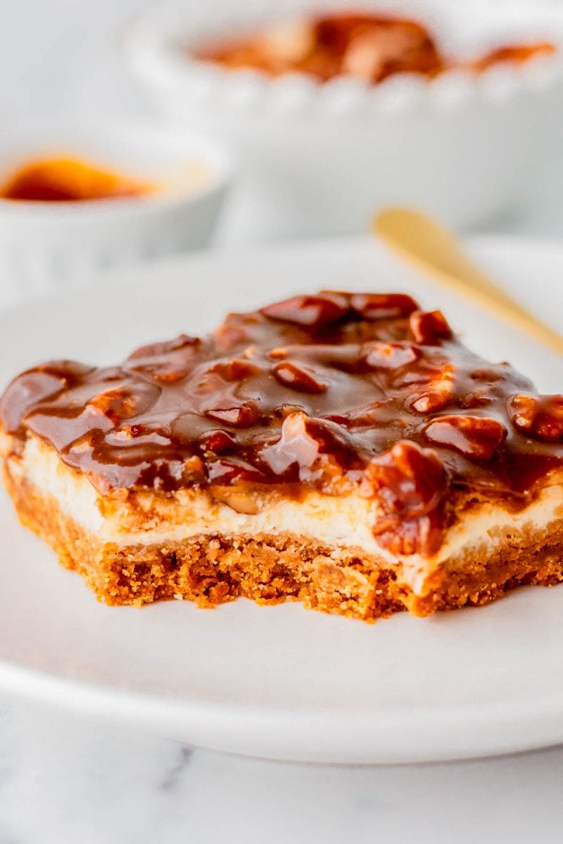 A square dessert bar with a crumbly base, creamy middle layer, and a glossy caramel nut topping is served on a white plate.