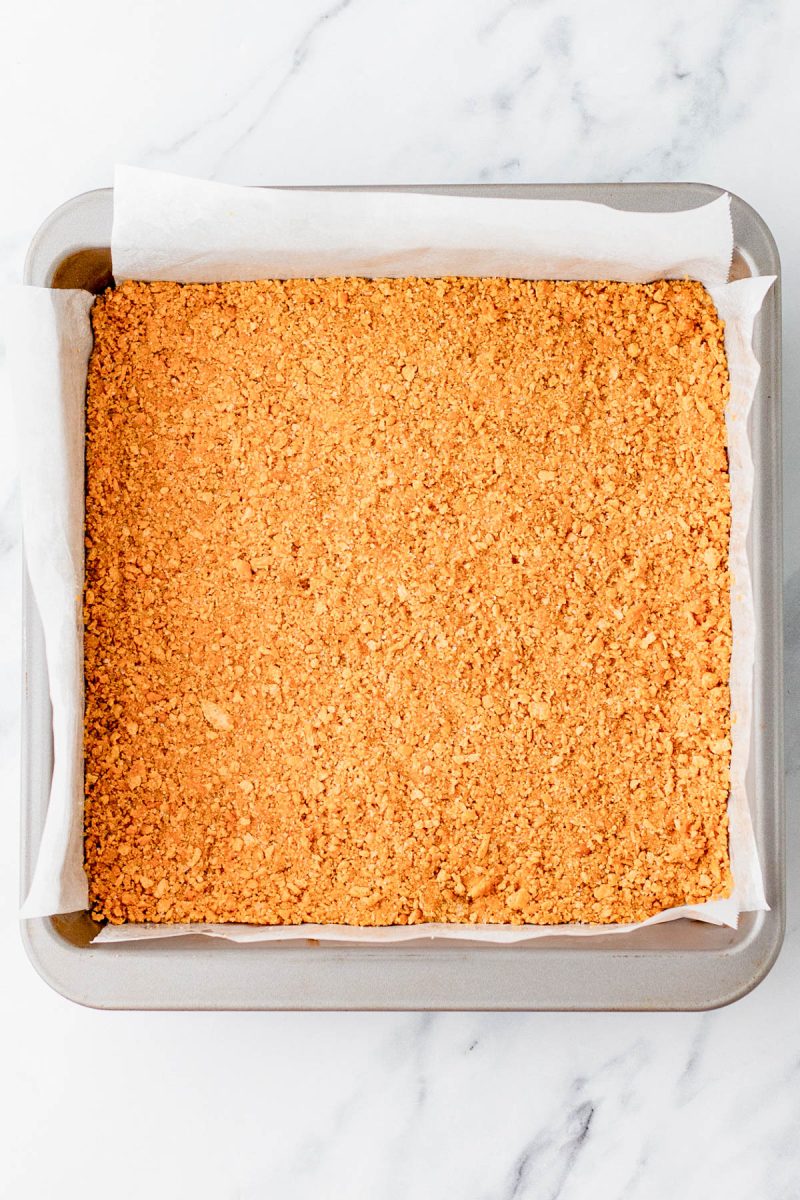 A square baking pan lined with parchment paper and filled with a pressed, crumbly golden crust sits on a marble surface.