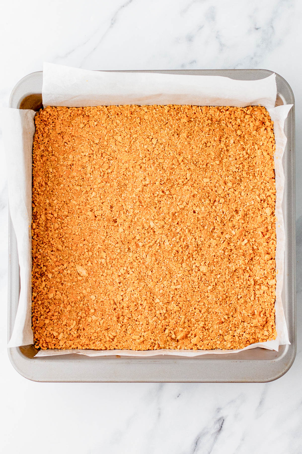 A square baking pan lined with parchment paper and filled with a pressed, crumbly golden crust sits on a marble surface.