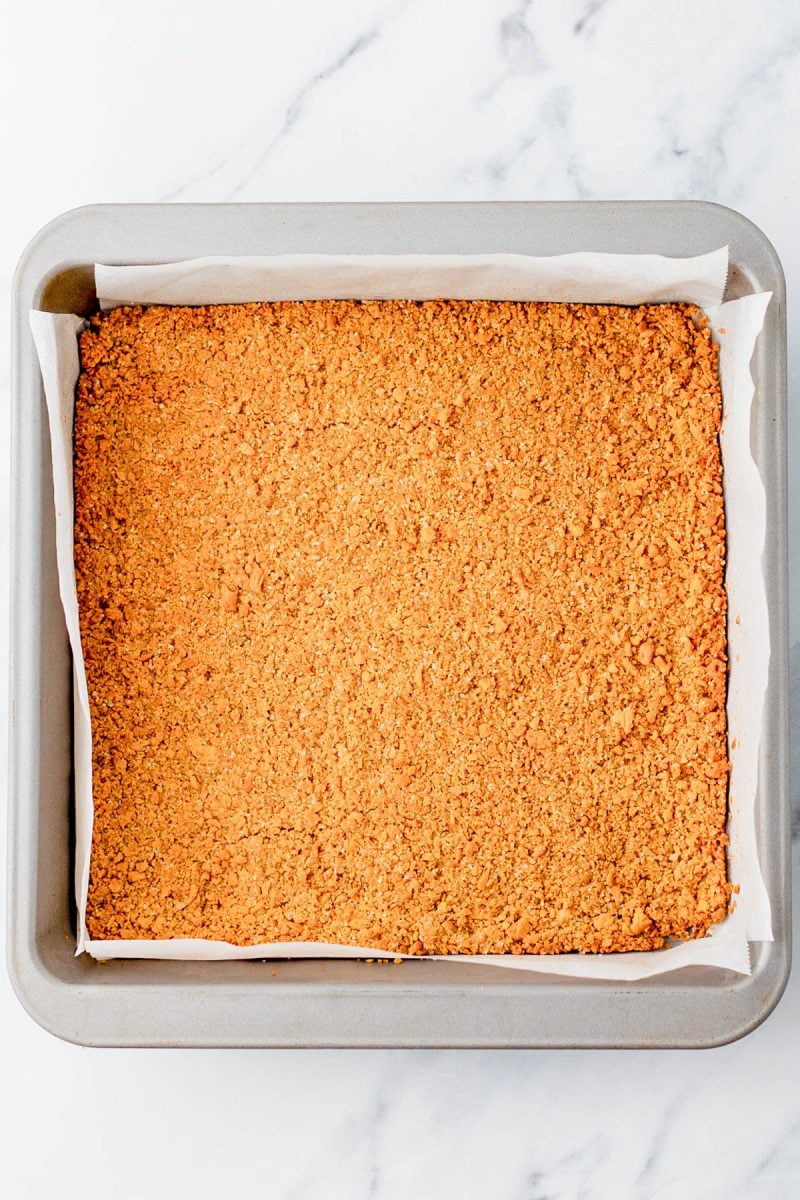 A square baking pan lined with parchment paper, filled with an even layer of crushed graham cracker crust on a white marble surface.