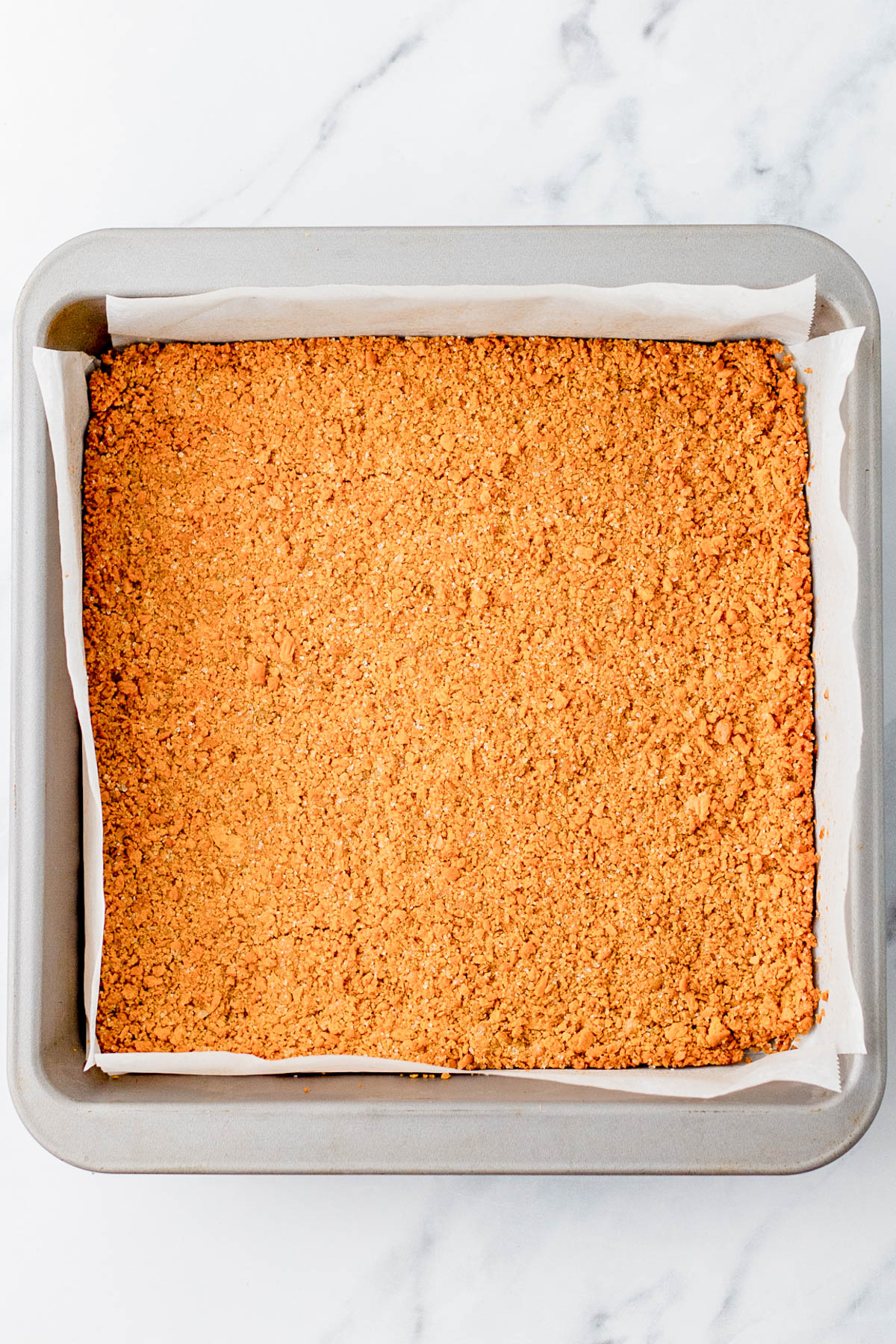A square baking pan lined with parchment paper, filled with an even layer of crushed graham cracker crust on a white marble surface.