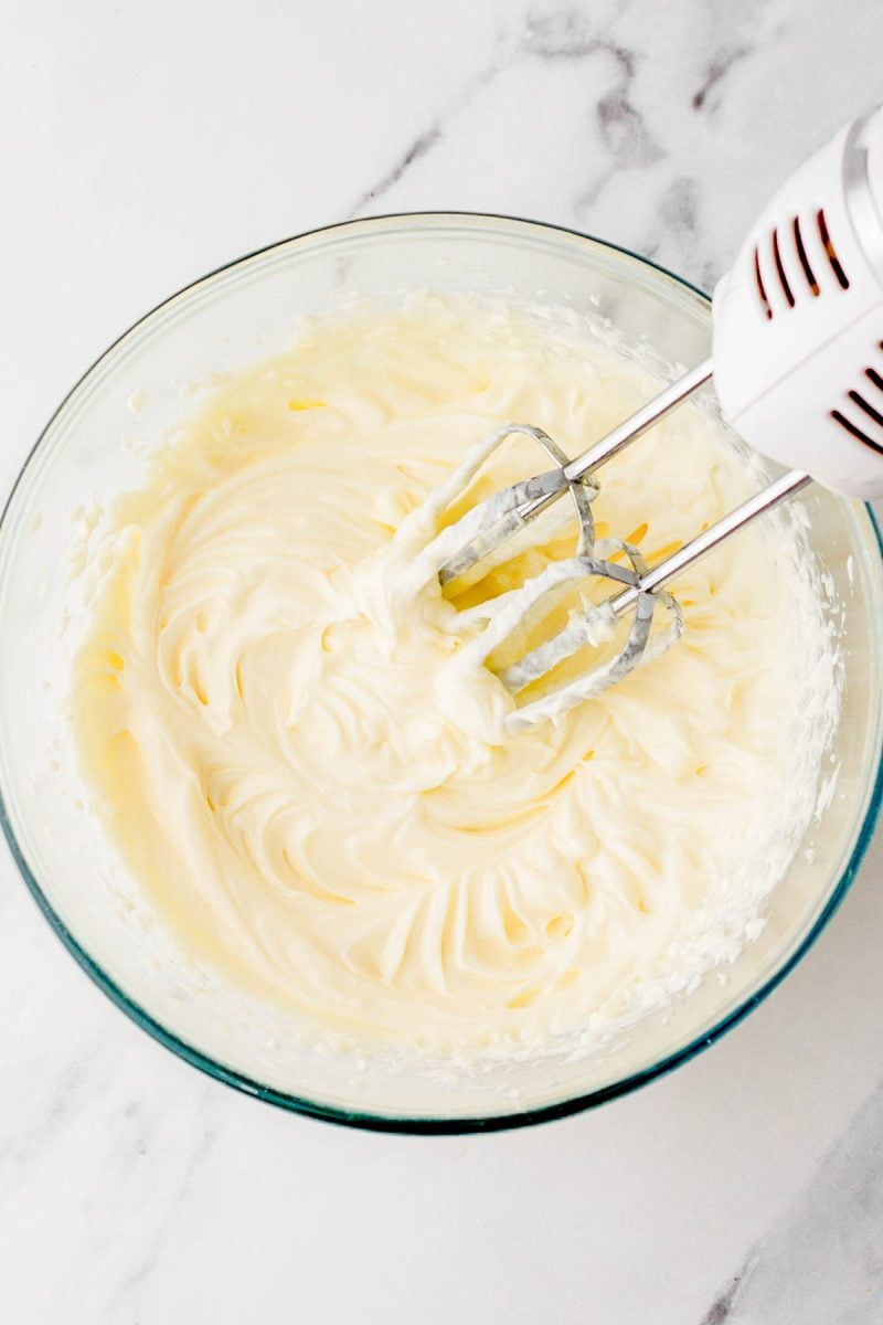 A glass bowl filled with creamy, whipped mixture being mixed by an electric hand mixer on a marble countertop.