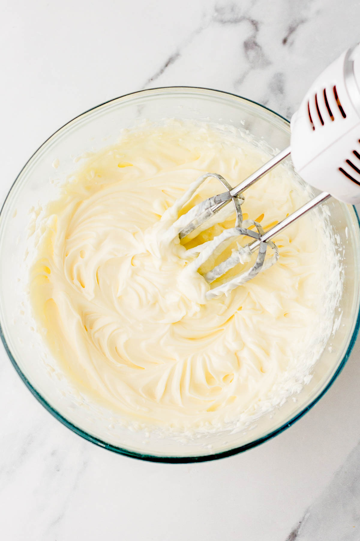 A glass bowl filled with creamy, whipped mixture being mixed by an electric hand mixer on a marble countertop.