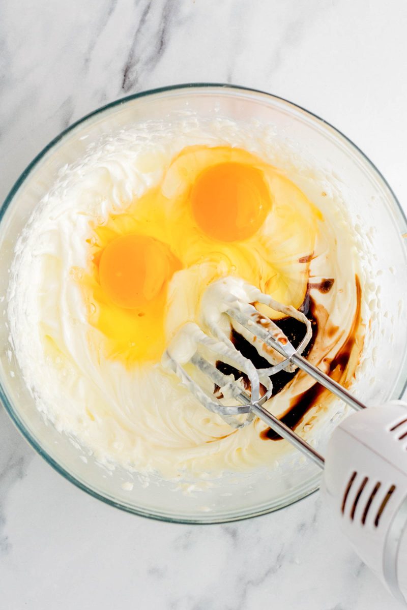 A glass bowl with creamed mixture, two cracked eggs, vanilla extract, and an electric hand mixer on a marble surface.