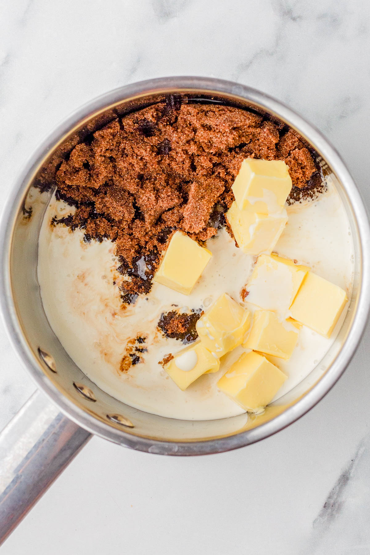 A saucepan containing brown sugar, cubed butter, and cream on a marble surface, ready to be mixed and heated.
