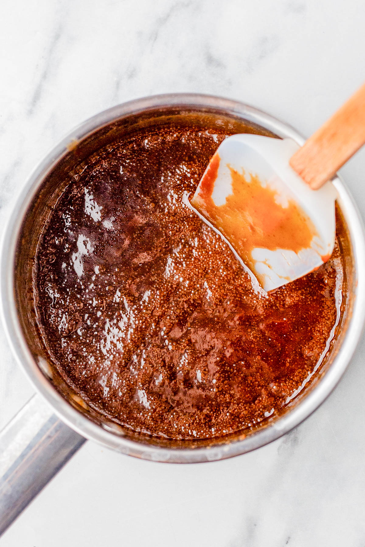 A saucepan filled with bubbling brown caramel sauce, being stirred with a white and wooden spatula on a marble surface.