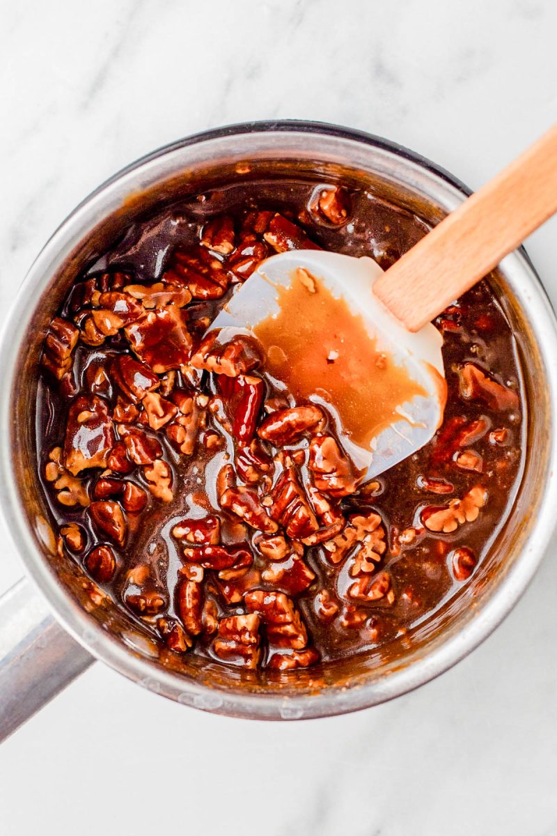 A saucepan filled with pecans coated in a thick brown caramel sauce, being stirred with a white spatula on a marble surface.