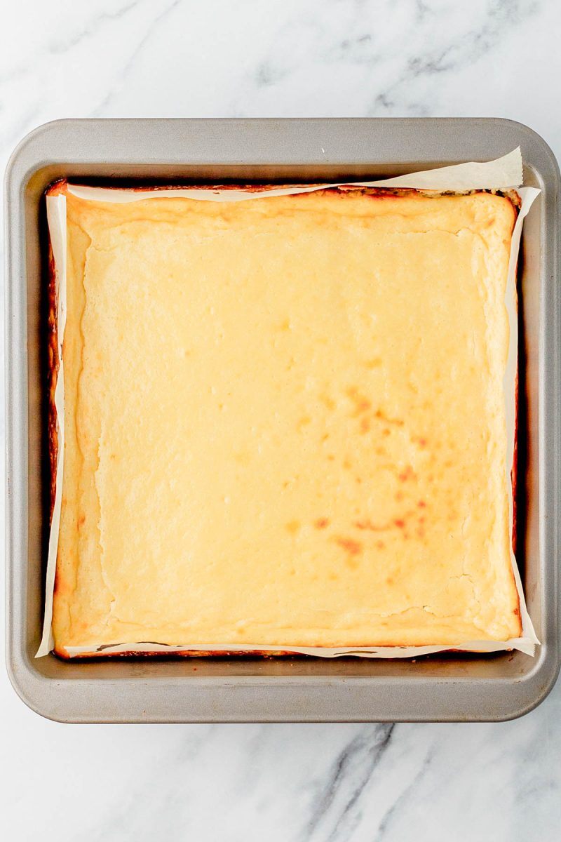 A baked cheesecake in a square pan lined with parchment paper, resting on a marble surface.