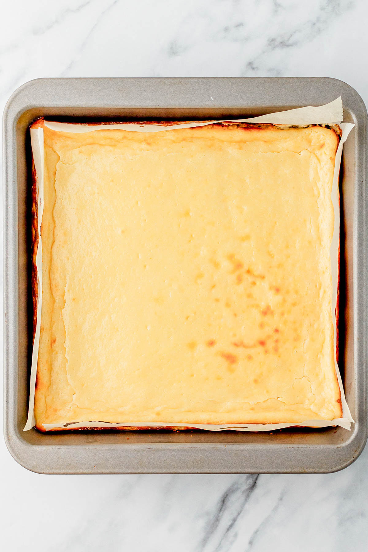 A baked cheesecake in a square pan lined with parchment paper, resting on a marble surface.