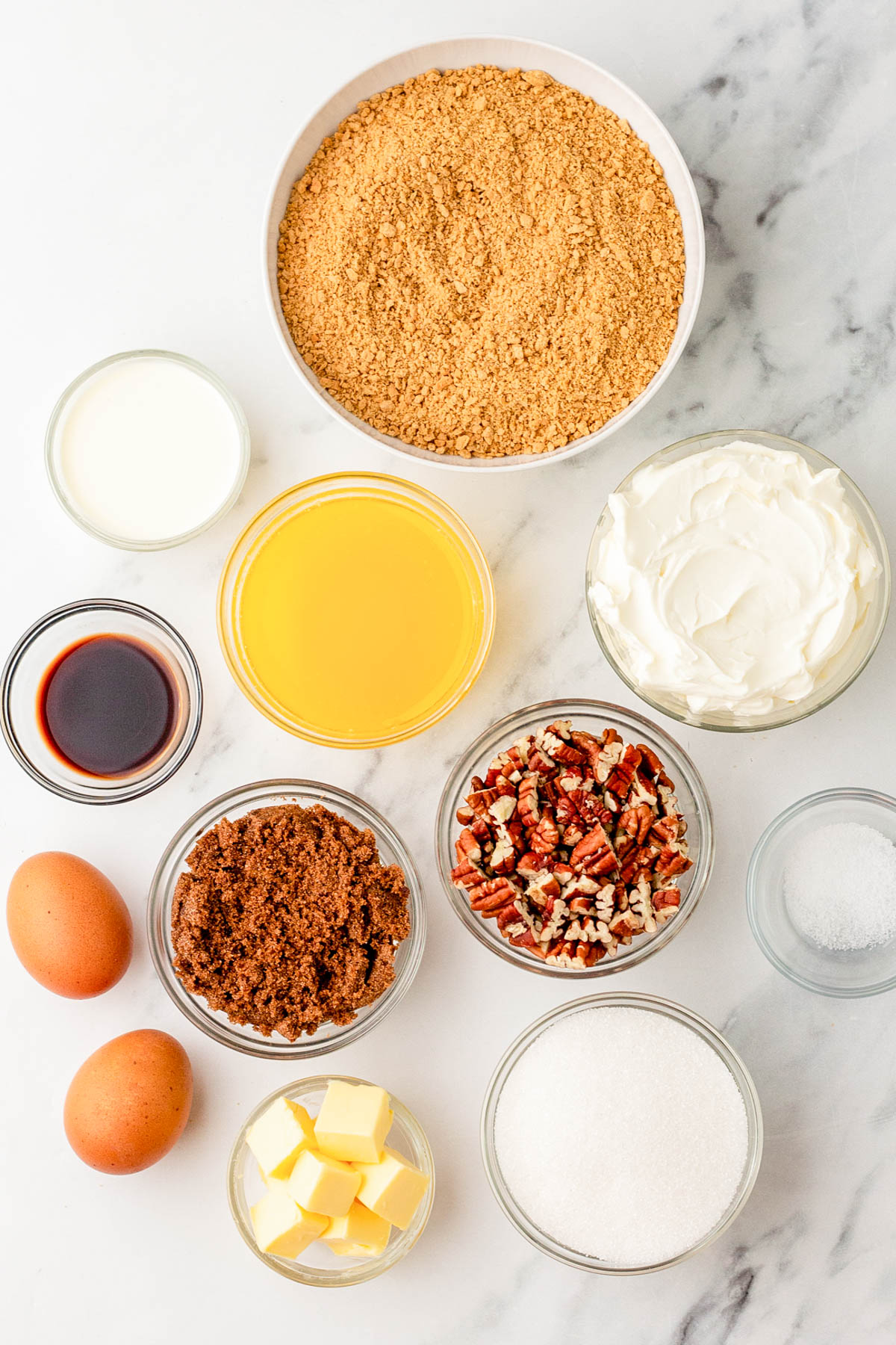 Overhead view of various cheesecake ingredients in bowls, including graham cracker crumbs, sugar, brown sugar, chopped pecans, eggs, butter, cream cheese, milk, vanilla, and salt.