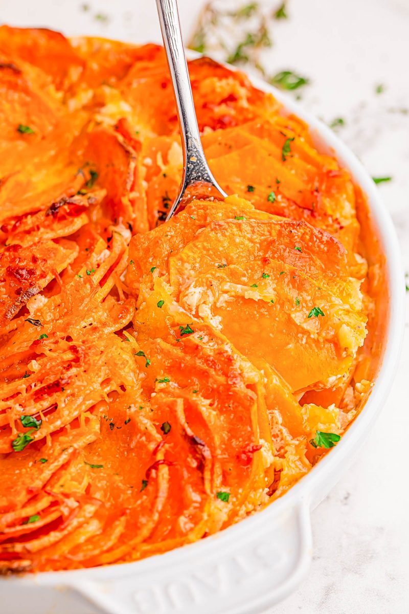 Golden, thinly sliced potatoes baked with cheese in a round white dish, with a serving spoon and a sprinkle of chopped herbs on top.