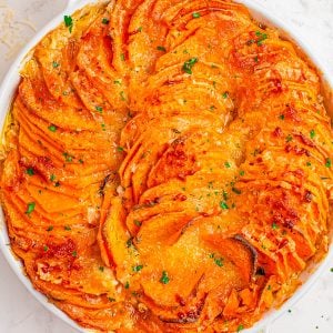 A baked sweet potato gratin in a round white dish, topped with melted cheese and garnished with chopped herbs.