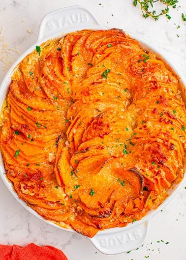 A baked sweet potato gratin in a round white dish, topped with melted cheese and garnished with chopped herbs.