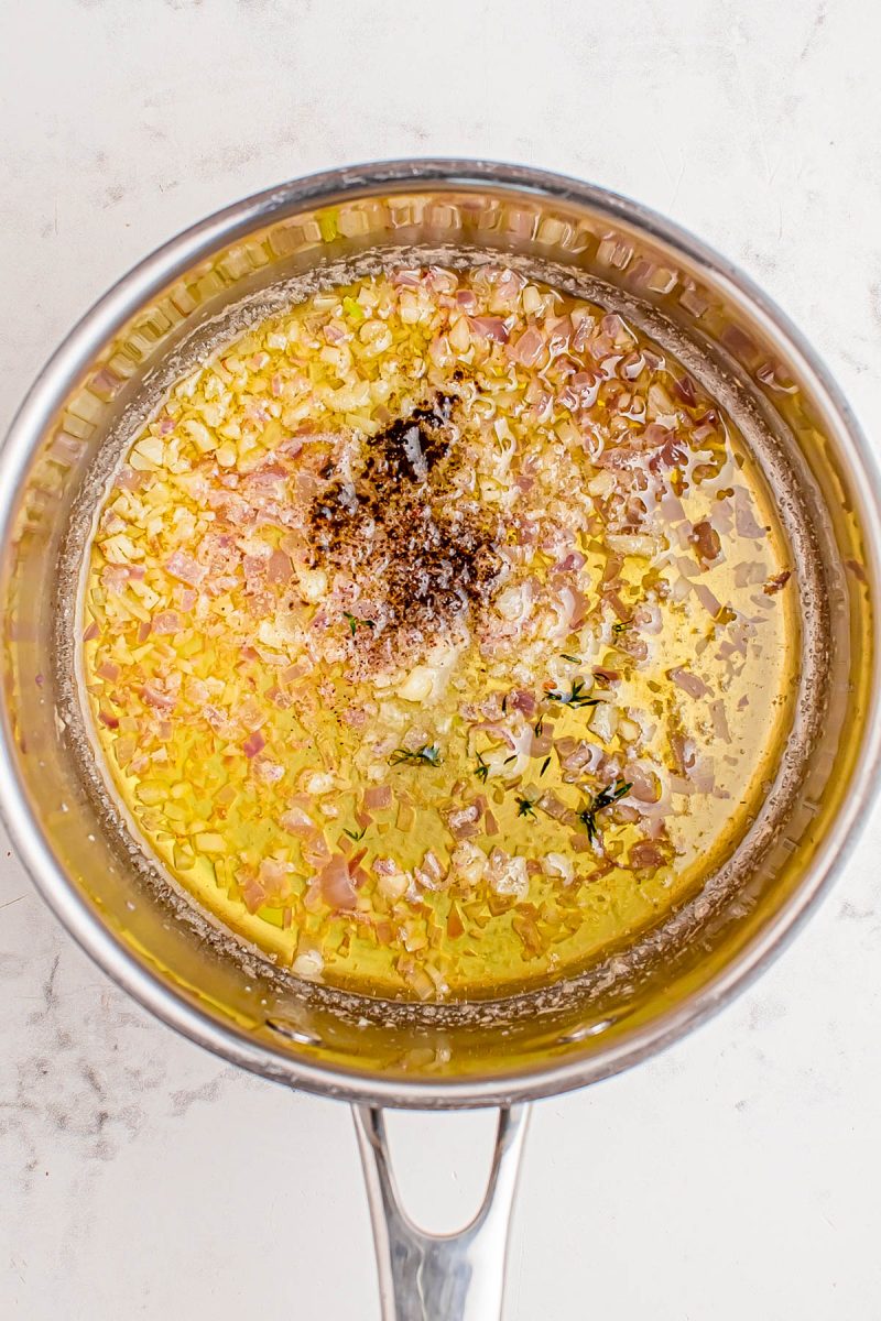 A saucepan containing melted butter, chopped shallots, herbs, and black pepper cooking together on a light countertop.
