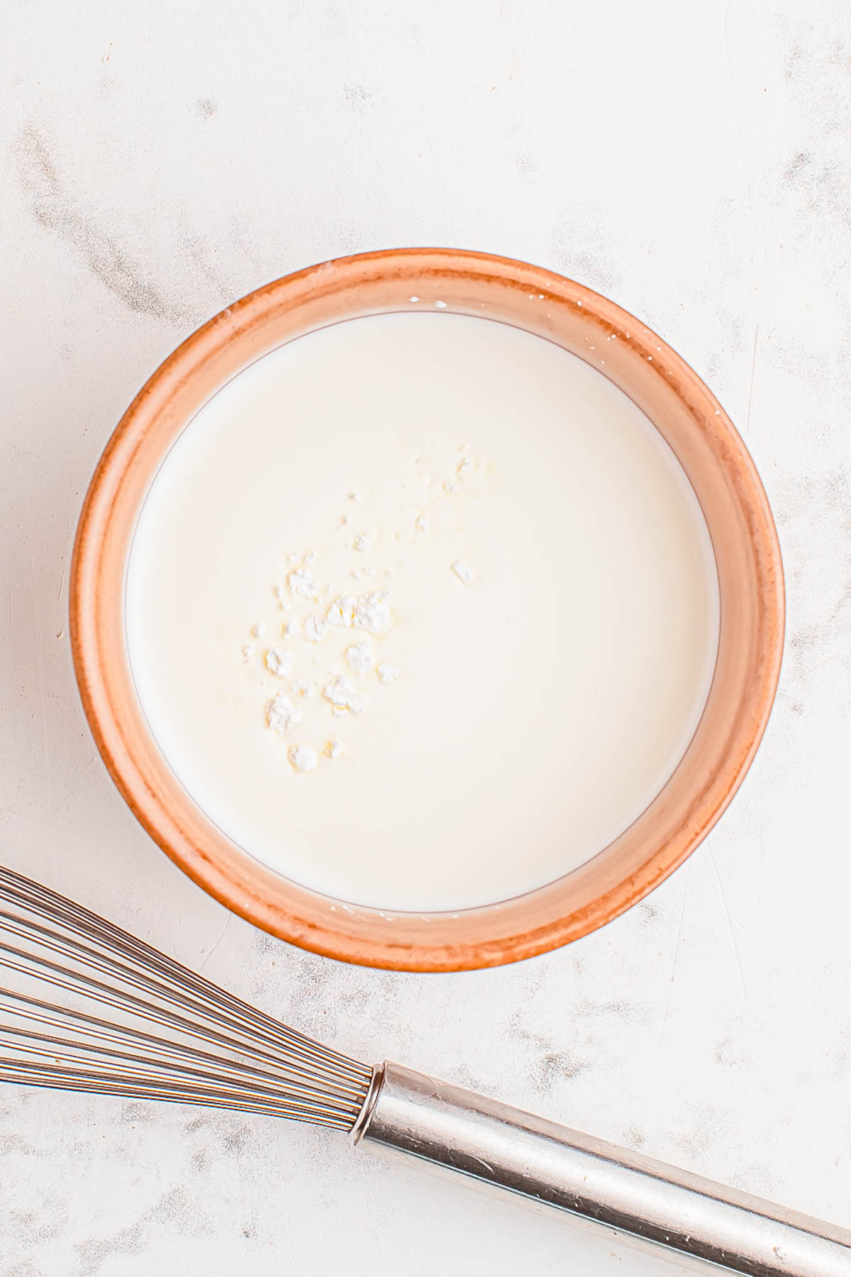 A bowl of milk with some white powder sprinkled on top stands next to a metal whisk on a white surface.