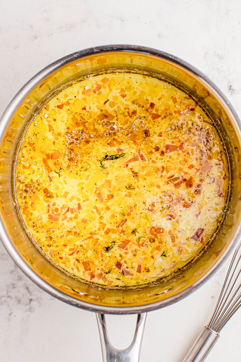 A saucepan containing a mixture of melted butter, oil, herbs, and finely chopped shallots or onions on a white countertop.