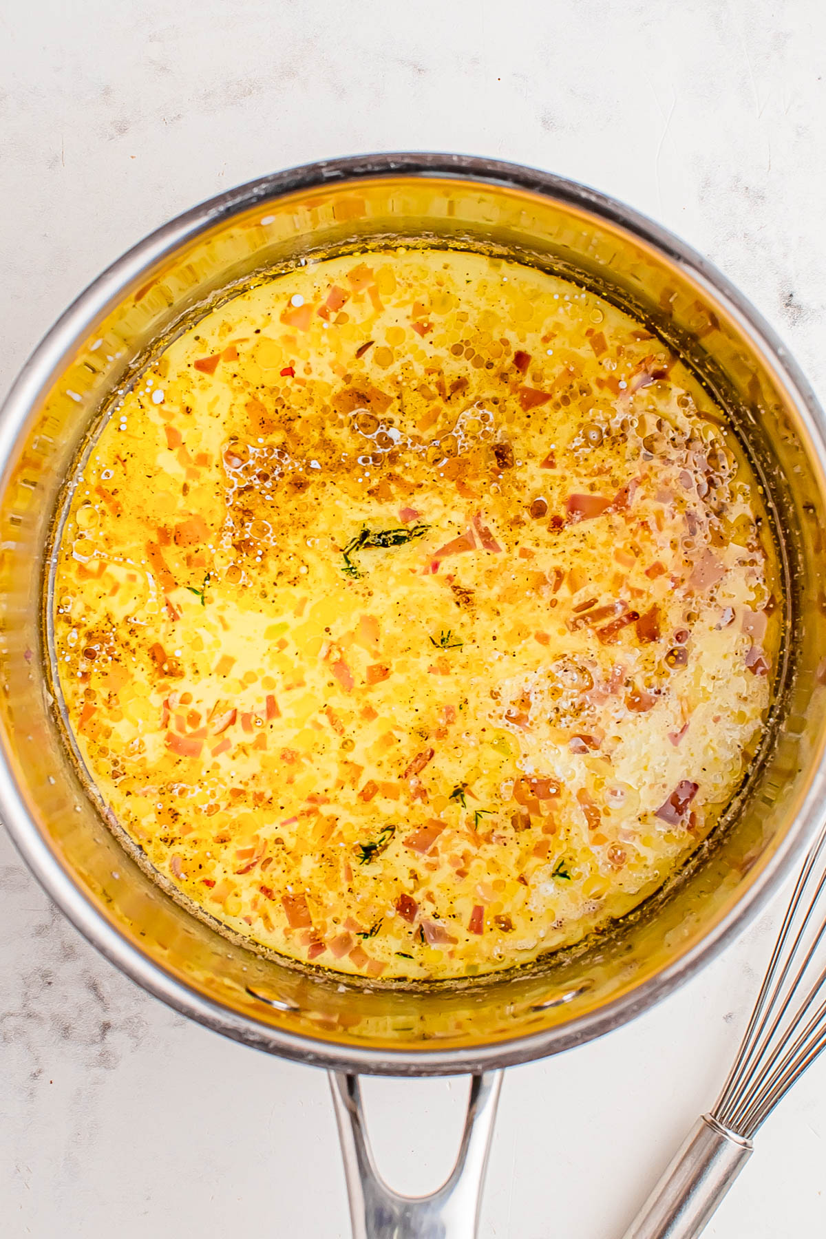 A saucepan containing a mixture of melted butter, oil, herbs, and finely chopped shallots or onions on a white countertop.