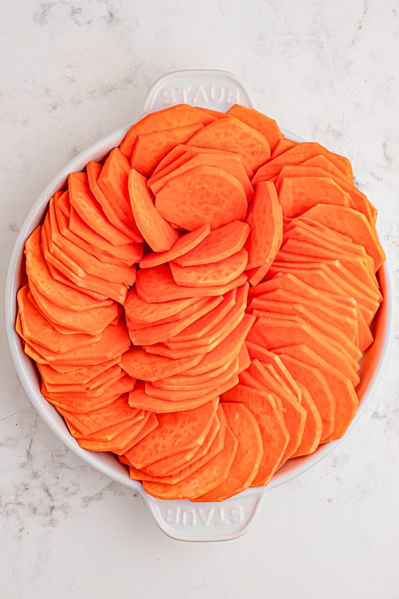 A round white dish filled with neatly arranged slices of raw sweet potato on a light marble surface.
