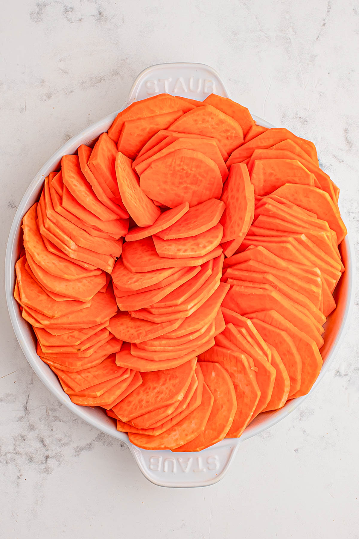 A round white dish filled with neatly arranged slices of raw sweet potato on a light marble surface.
