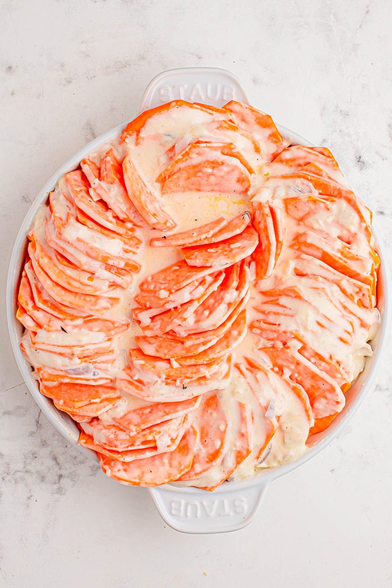A round white baking dish filled with thinly sliced sweet potatoes arranged in a spiral pattern, covered with melted cheese on top.