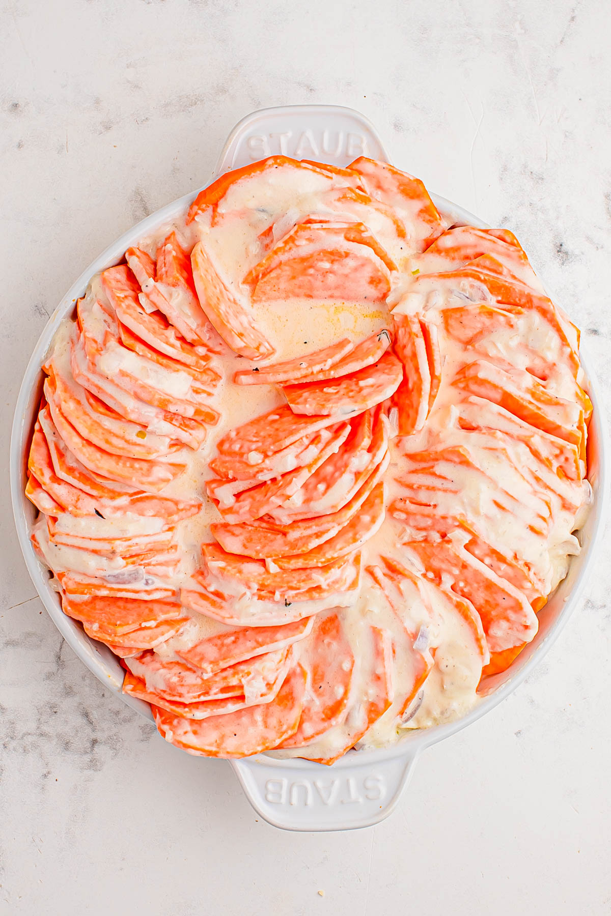 A round white baking dish filled with thinly sliced sweet potatoes arranged in a spiral pattern, covered with melted cheese on top.