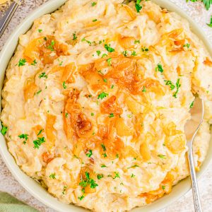 A bowl of creamy mashed potatoes topped with caramelized onions and chopped parsley, with a spoon resting inside.