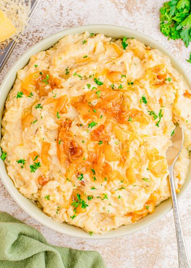 A bowl of creamy mashed potatoes topped with caramelized onions and chopped parsley, with a spoon resting inside.