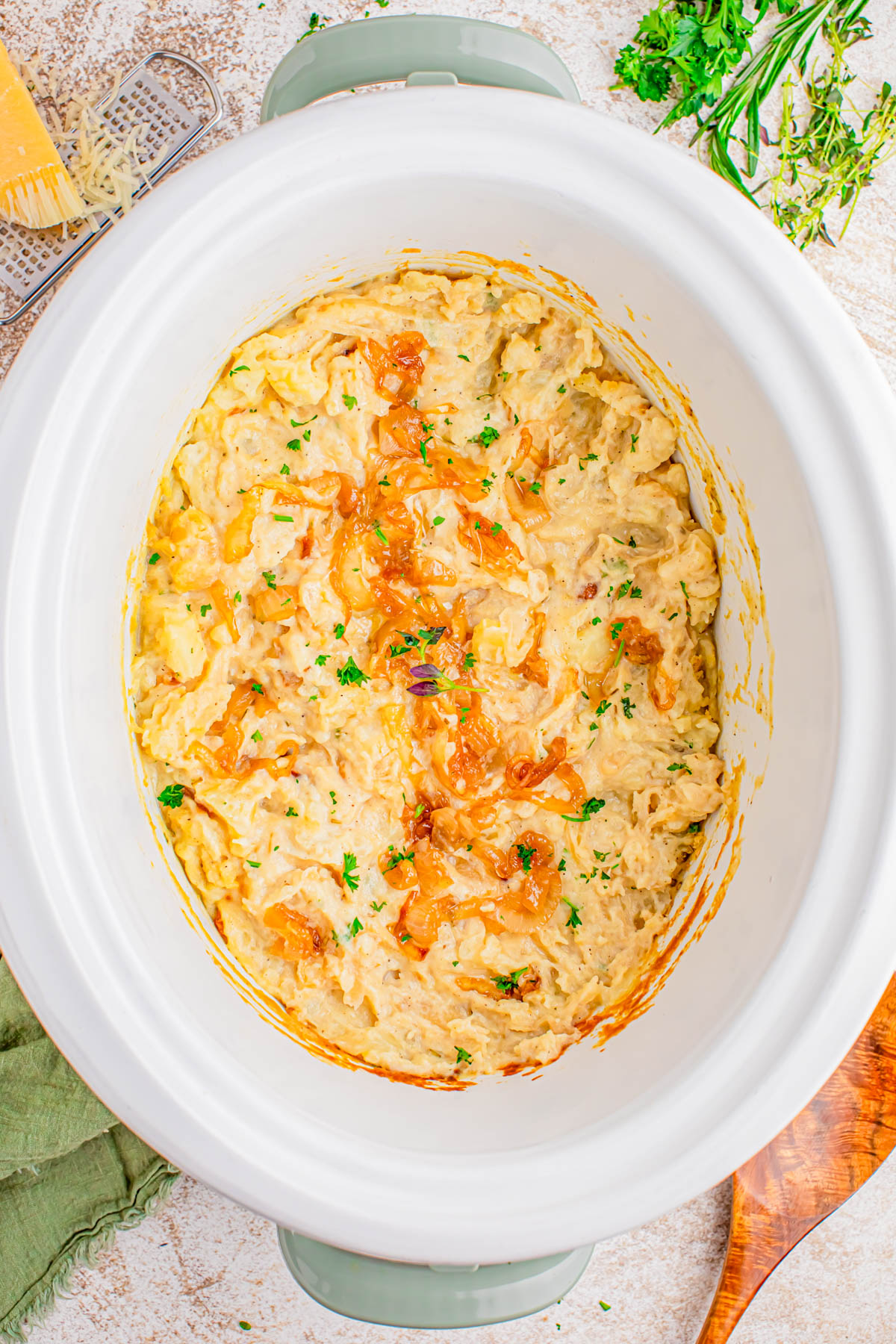 A slow cooker filled with creamy macaroni and cheese, topped with caramelized onions and chopped parsley, sits on a light-colored countertop.