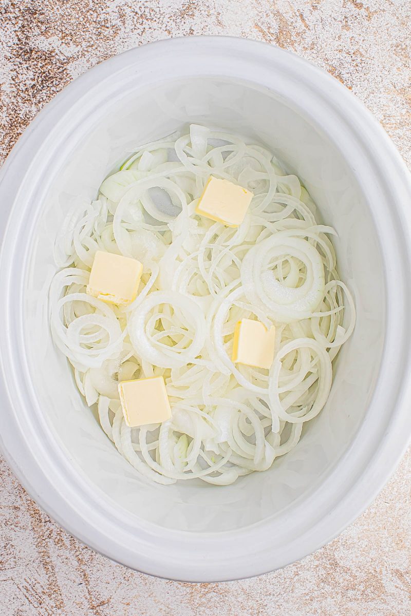 Sliced onions and four cubes of butter placed inside a white slow cooker on a light textured surface.