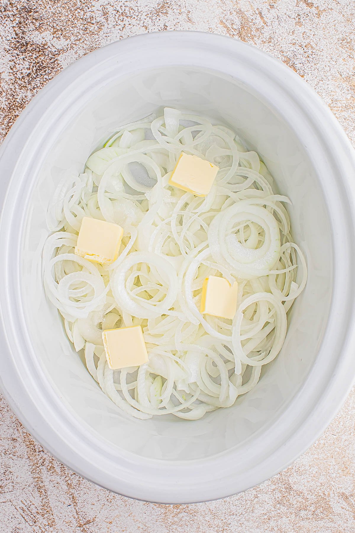 Sliced onions and four cubes of butter placed inside a white slow cooker on a light textured surface.