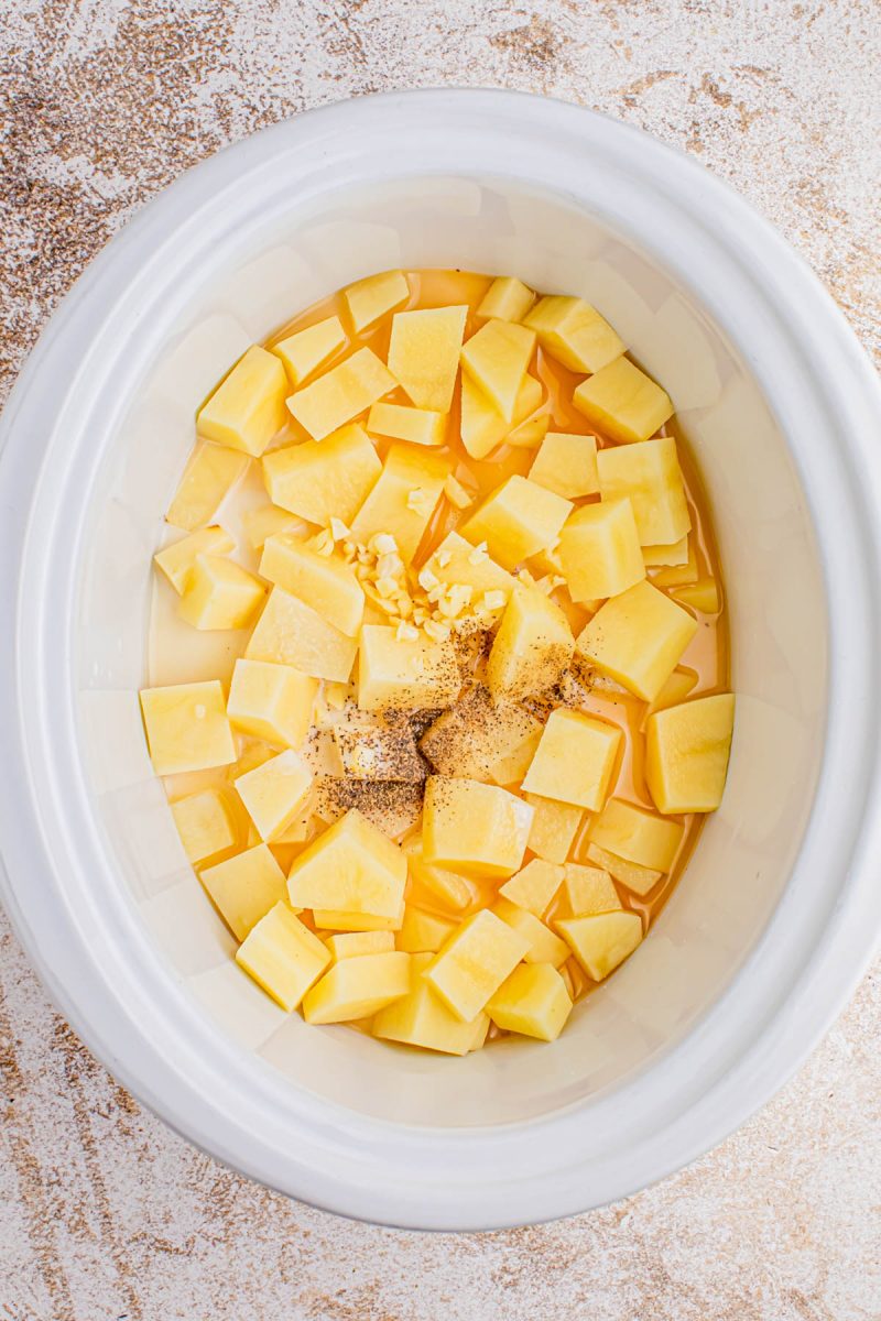 Diced potatoes, minced garlic, black pepper, and liquid are in a white slow cooker on a light textured surface.