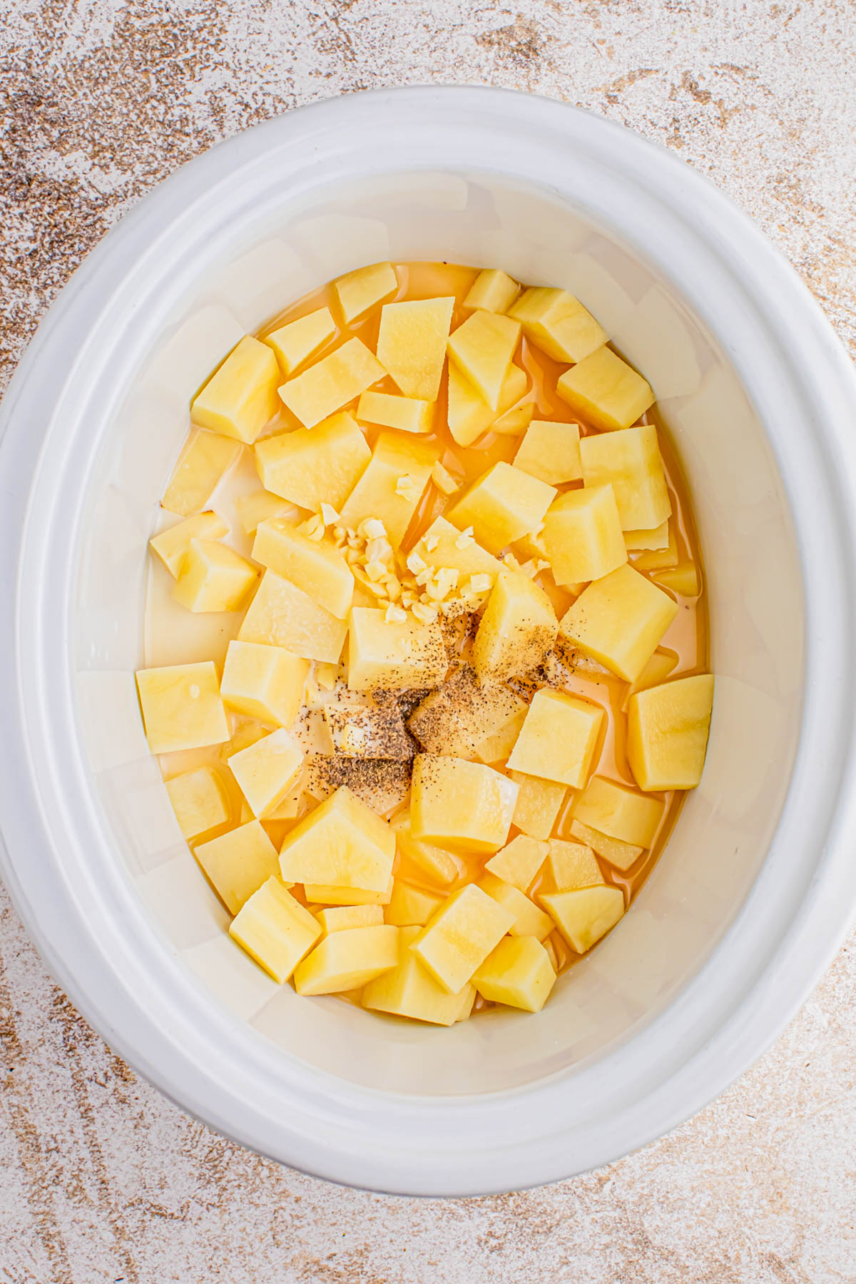 Diced potatoes, minced garlic, black pepper, and liquid are in a white slow cooker on a light textured surface.