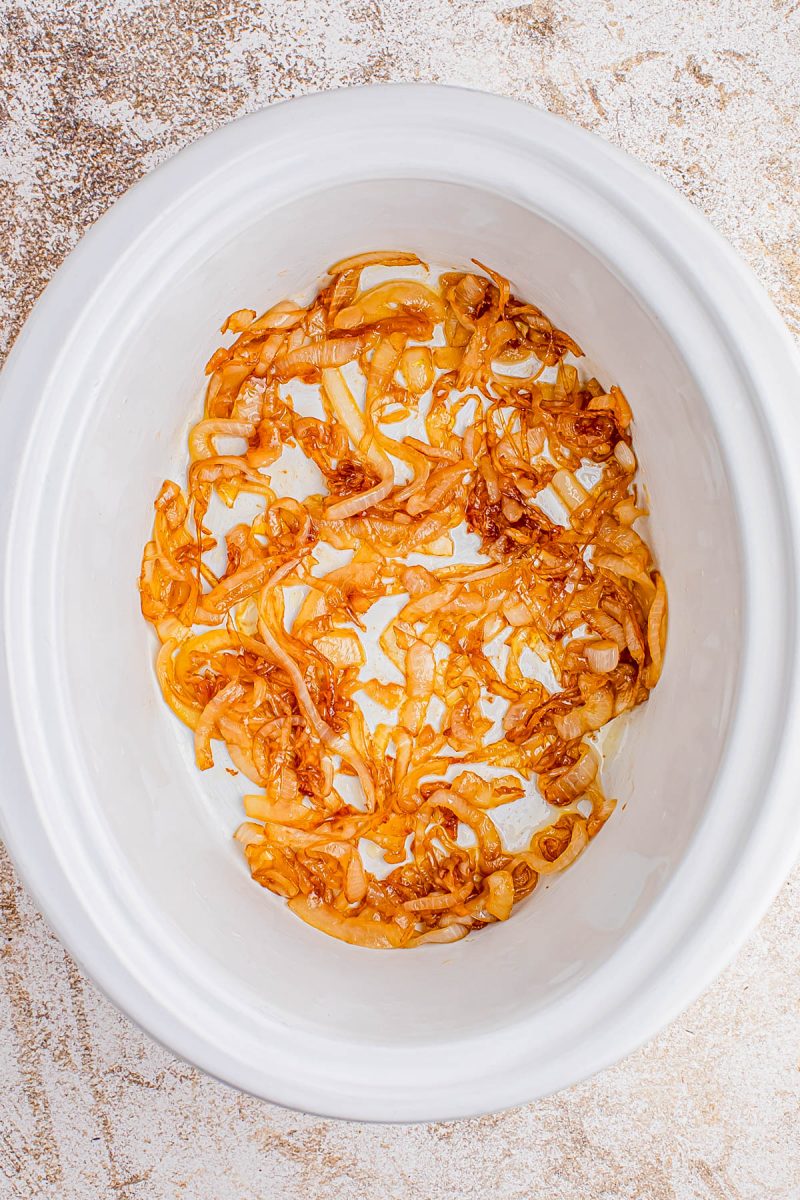 Sliced caramelized onions spread at the bottom of a white slow cooker on a light textured surface.