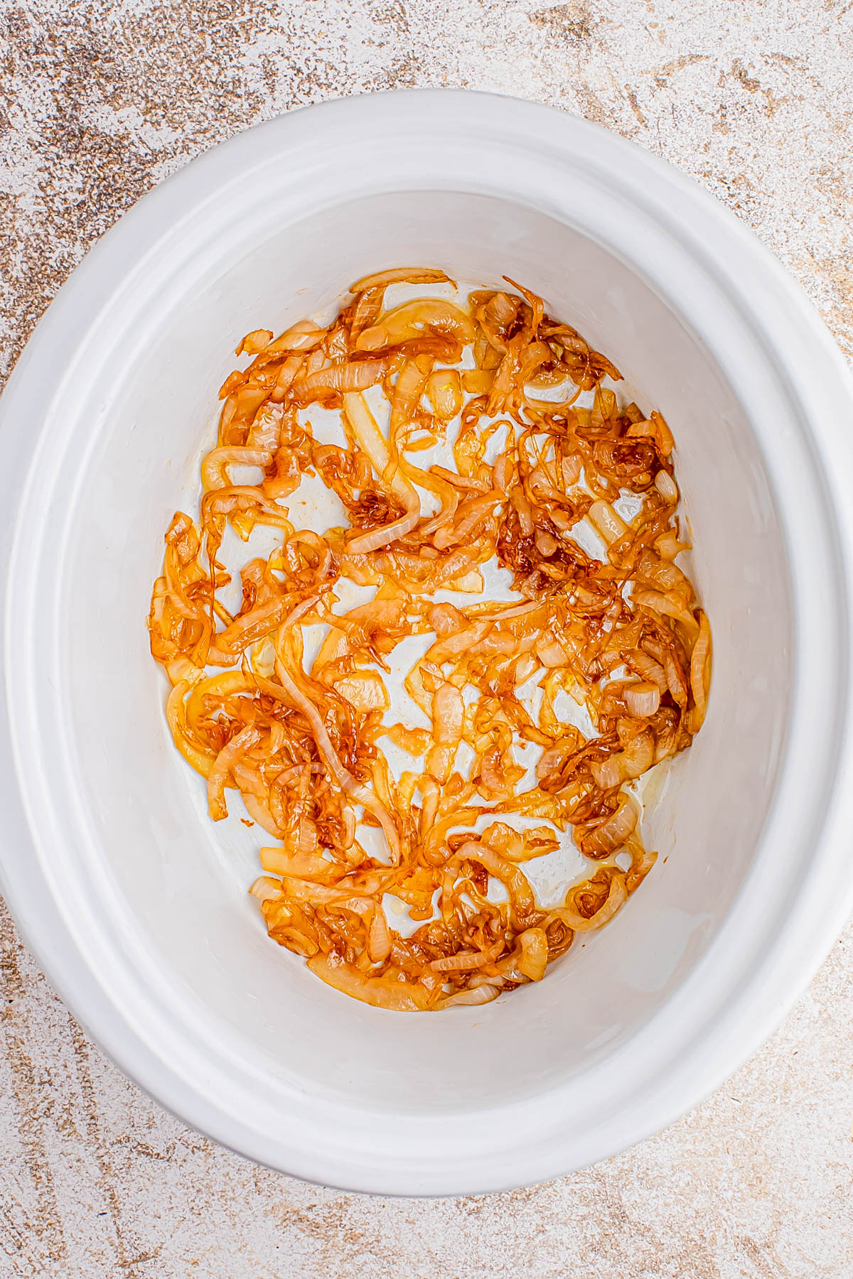 Sliced caramelized onions spread at the bottom of a white slow cooker on a light textured surface.