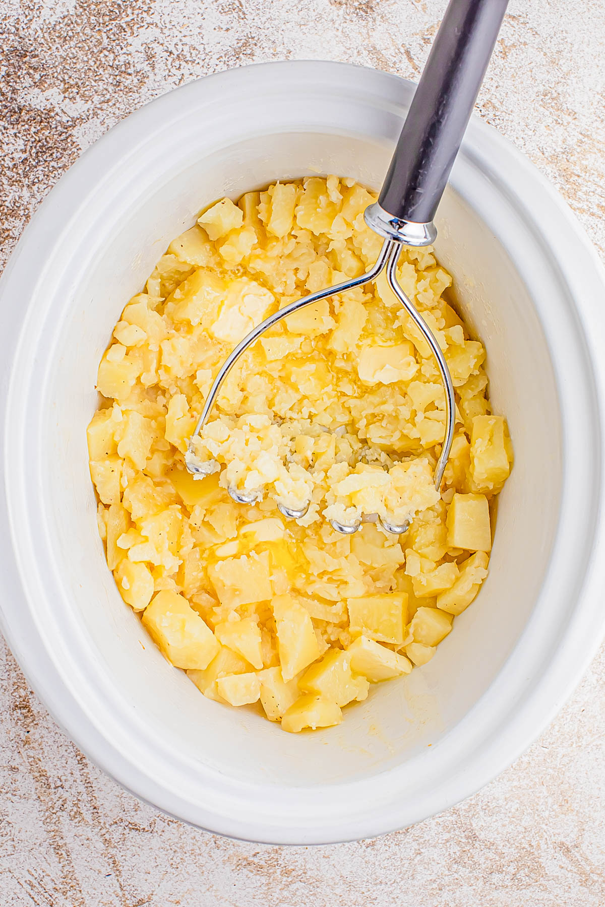 Cubed potatoes being mashed in a white slow cooker with a metal potato masher.