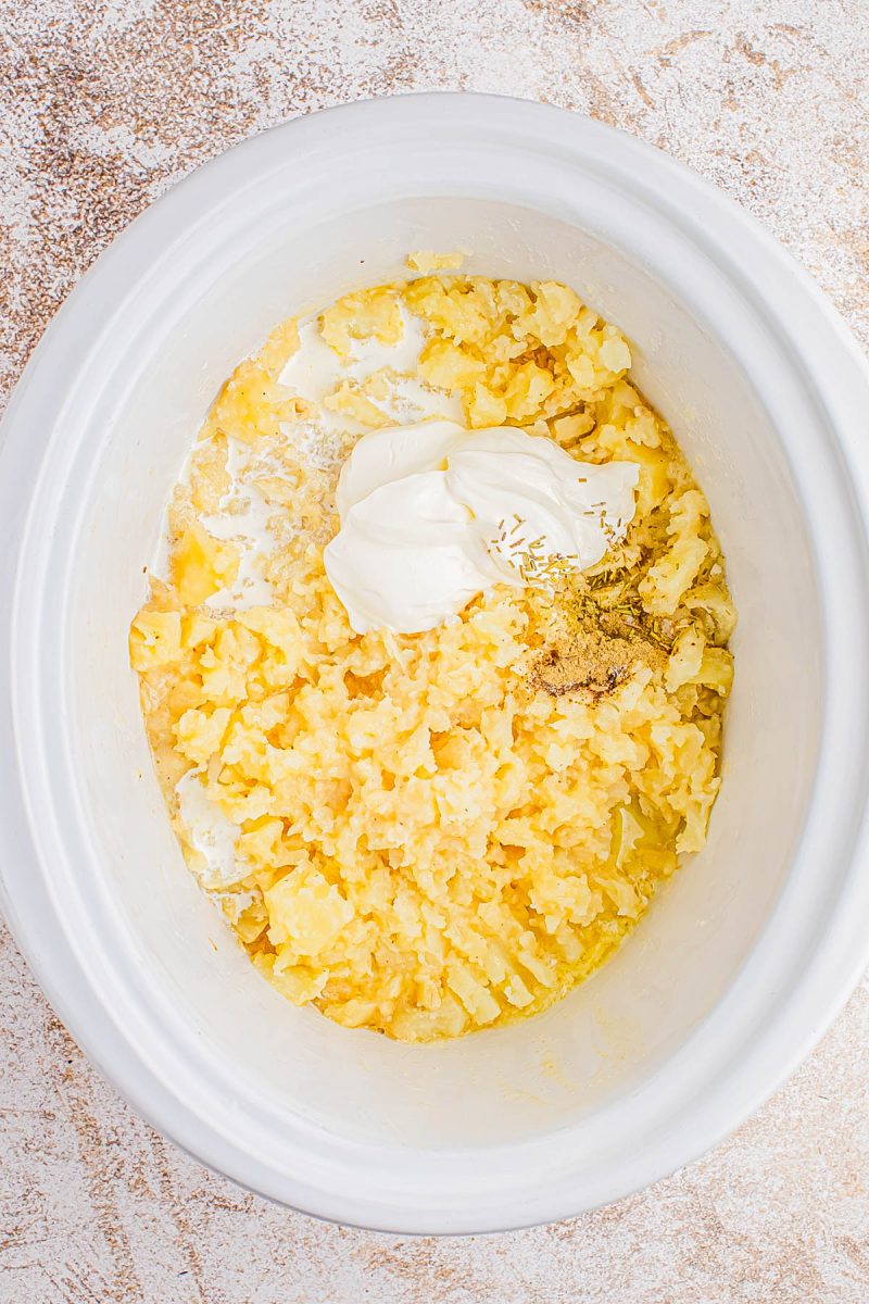 Mashed potatoes with butter, sour cream, and seasonings in a white slow cooker on a textured countertop.