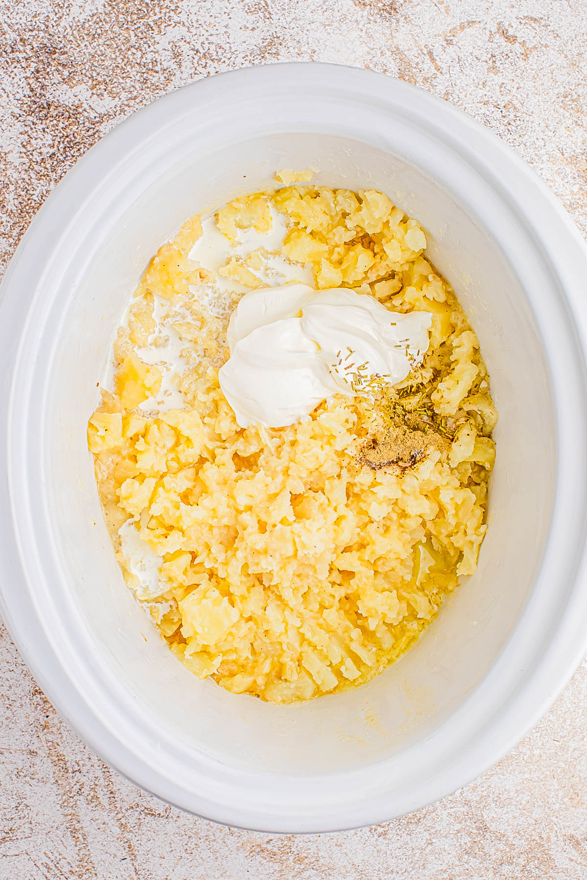 Mashed potatoes with butter, sour cream, and seasonings in a white slow cooker on a textured countertop.