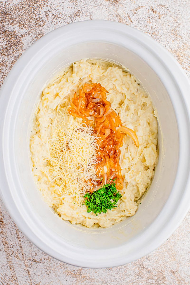 A slow cooker filled with a creamy potato mixture, topped with caramelized onions, shredded cheese, and chopped parsley.