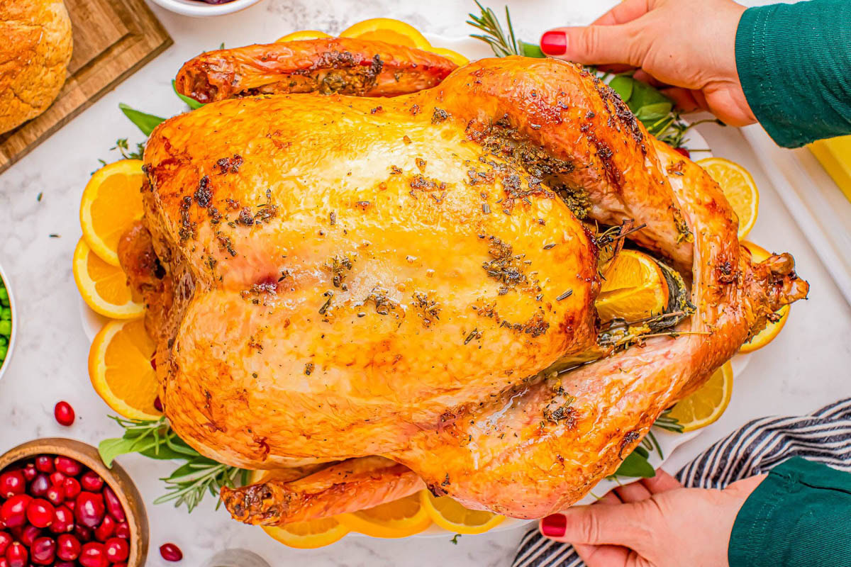 A whole roasted turkey garnished with herbs and orange slices is placed on a platter, with hands preparing to serve it. Bowls of cranberries and vegetables are nearby.