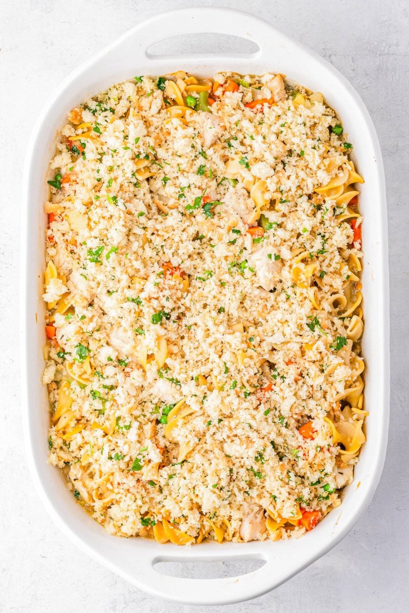 A white baking dish filled with a casserole topped with a crumbly mixture, containing visible pasta, vegetables, and herbs.